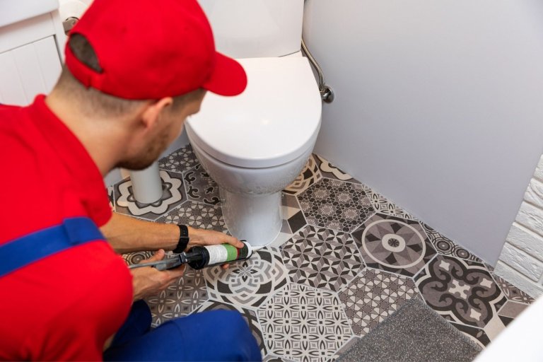 Plumber in red and blue uniform caulks around base of toilet.